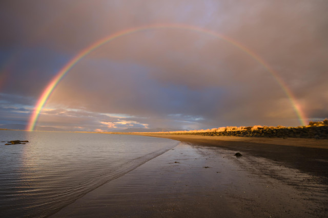 A picture of a large bright rainbow seen from a black sandy beach in Iceland.