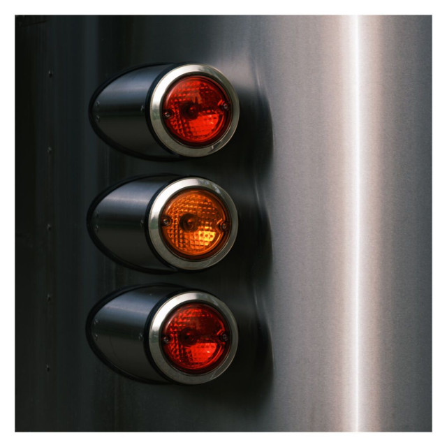 This is a square color image showing three lights mounted vertically on a metallic surface. The lights are cylindrical and extend outward from the surface, each enclosed in a metal housing with circular lenses. The top and bottom lights are red, while the middle one is orange. The metallic surface on which the lights are mounted has a smooth, reflective finish with soft lighting, which creates a slight gradient effect from left to right. The texture of the metal and the glossy finish of the lights create a sleek and industrial appearance. The overall composition is minimalist, with the lights adding a pop of color against the metallic background.