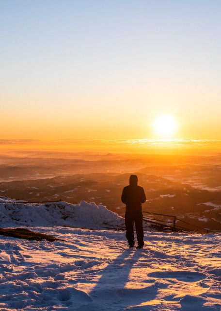 Blick von einem Berg auf eine Hügel- und Tallandschaft. Es liegt Schnee und die Silhouette eines Menschen ist im Vordergrund. Am Horizont sieht man die gerade aufgegangene Sonne. Es gibt ganz wenige Wolken.