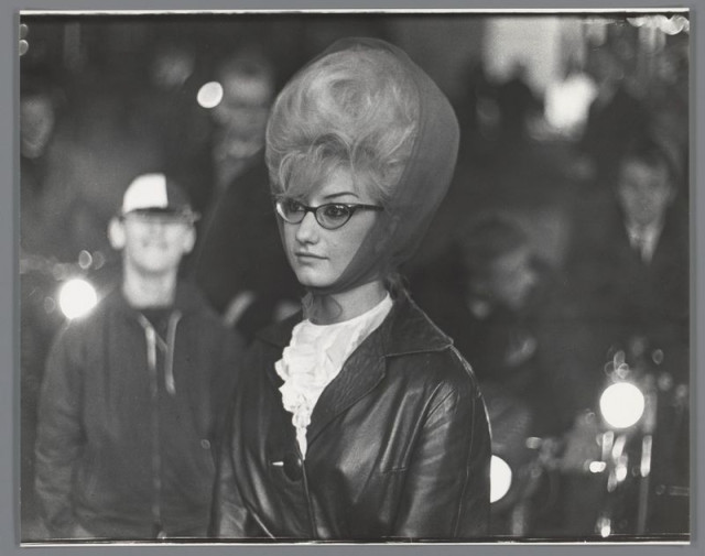 The image is a black and white photograph featuring a woman in the foreground with an unusual, voluminous hairstyle. She wears glasses and has on what appears to be a leather jacket over a ruffled blouse or dress. The background shows blurred figures of other people, suggesting that she may be at a crowded event or public gathering. There are lights visible which could indicate nighttime setting, possibly outdoors given the bokeh effect typical for photography taken in low light conditions with fast shutter speeds. This scene reflects street photography commonly found during the mid-20th century and is associated with Ed van der Elsken's work captured at events like fairs or carnivals in Amsterdam as noted by the referenced source.