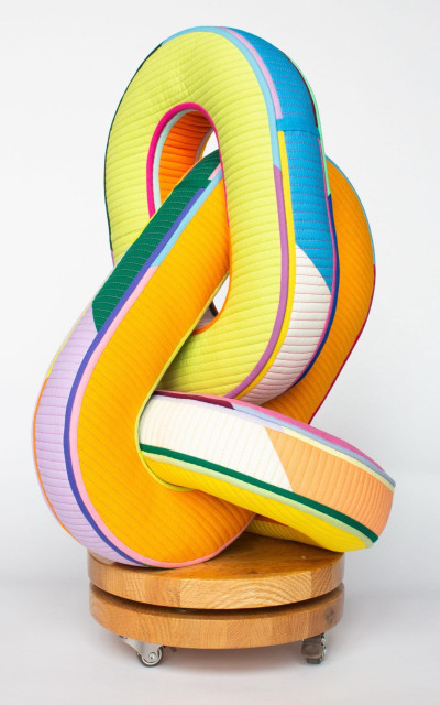 a sculptural textile piece resembling three chain links with different colored facets, perched on a wooden base with wheels