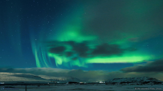 A photo taken at night, with many stars visible above a heavy grey cloud bank on the horizon. Below it, scattered lights of buildings dot the snow-covered ground. High in the sky is a gorgeous green block of light, partially hidden behind clouds. It twists and swirls in streaks and shafts, some almost vertical, some almost horizontal. At the lower edge, the green contains a hint of red and yellow.