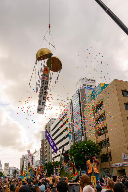 A colorful parade scene featuring two large, hanging golden ornaments with ribbons, surrounded by a shower of small, colorful balls. A crowd of spectators captures the moment with their phones, while buildings and banners are visible in the background, suggesting a festive atmosphere.