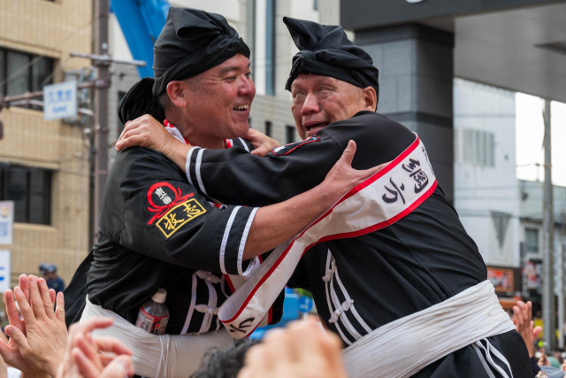 Two men in traditional black outfits and head wraps are joyfully embracing during a celebration. They appear to be participating in a festive event, with onlookers cheering in the background.