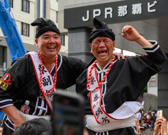 Two men in traditional black attire with red and white accessories are joyfully celebrating at a public event. One appears to be laughing while the other is expressing intense emotion. The background features a sign for "JPR" and a crowd of spectators.