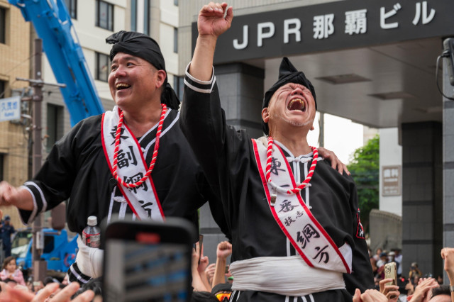 Two men in traditional Japanese clothing celebrate joyfully, raising their arms and smiling. They wear black outfits with white sashes and red beads, amidst a lively crowd. The background features a modern building with the letters "JPR."