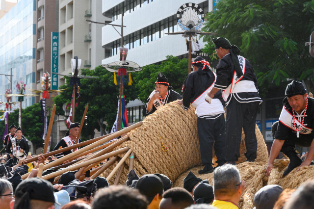 A lively festival scene featuring men in traditional attire working together to lift large bundles of rope. The background includes crowds of onlookers, buildings, and decorative banners, creating a vibrant atmosphere.