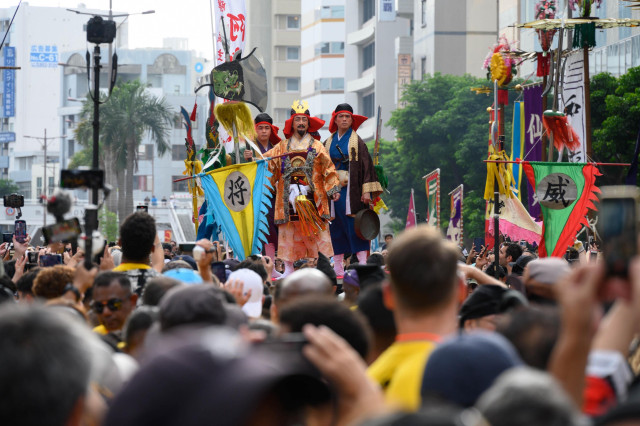 A vibrant parade scene with performers in traditional attire holding colorful flags and banners. The central figure is adorned in elaborate clothing, while a large crowd captures the moment with cameras and smartphones. Buildings and greenery frame the background, indicating an urban setting.