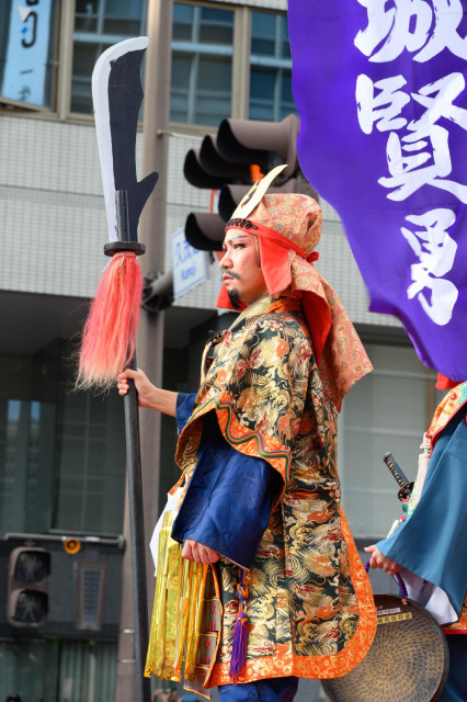 A man dressed in traditional ornate clothing holds a staff with a distinctive blade and a colorful tassel. He has a stern expression and is participating in a festival or parade, accompanied by another figure in the background. A purple banner with white text is visible