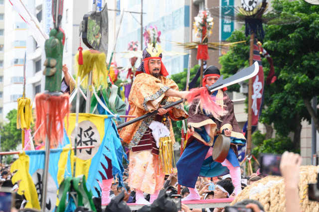 A vibrant parade scene featuring performers in traditional costumes wielding swords and other props. Colorful banners and decorations surround them, with a crowd of spectators in the foreground. The setting appears lively and festive.