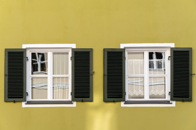 Gelbe Hauswand, zwei Fenster nebeneinander, jeweils vier Scheiben. 
In je einer Scheibe spiegelt sich ein ggü liegendes Fenster.
Die beiden Fenster haben Läden und weiße Umrandungen.