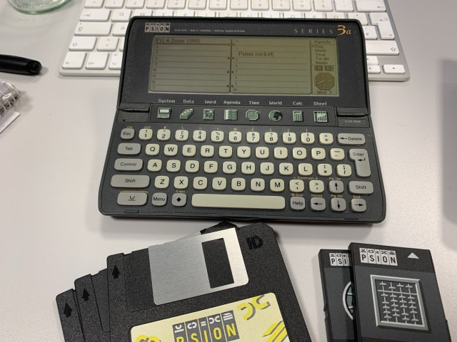 A vintage Psion Series 3a handheld computer is displayed on a desk. It features a keyboard and a screen showing a dated agenda. Nearby, several Psion data cartridges are visible. A white keyboard and a pen are also present in the background