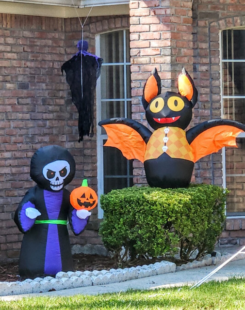 Inflatable Halloween decorations near the entrance of a home. An orange and black bat, a cloaked skeleton with a pumpkin, and a ghost.