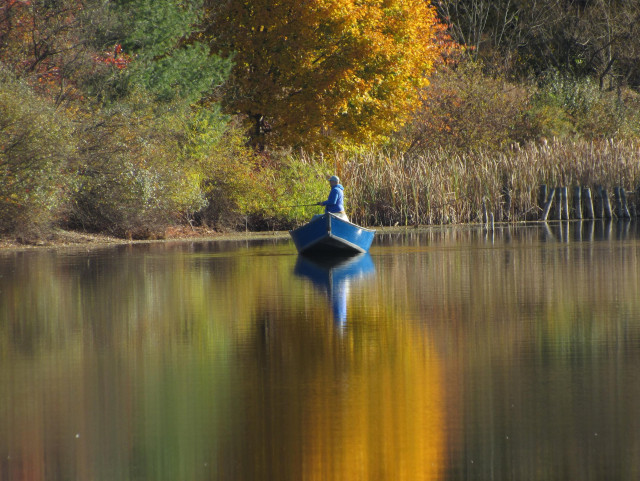 A fisherman tries his luck along the opposite shore. He is in a blue boat and wearing a blue jacket which is reflected on the surface of the water.