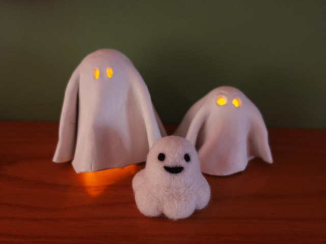 A group of three white ghosts on a mantelpiece. The back two are made from air dry clay, one taller than the other and their eyes are lit up by a tealight inside. The one in front is Sebastian, a smiling needlefelted little squidge of a ghost.