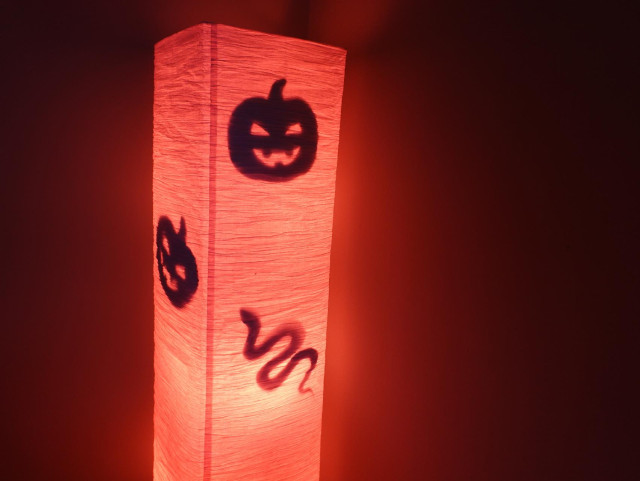 A tall floor lamp with simple cardboard cutouts of pumpkins and a snake stuck to the inside of the shade and visible in silhouette. The bulbs have been changed to red, casting an eerie glow around the surrounding walls.