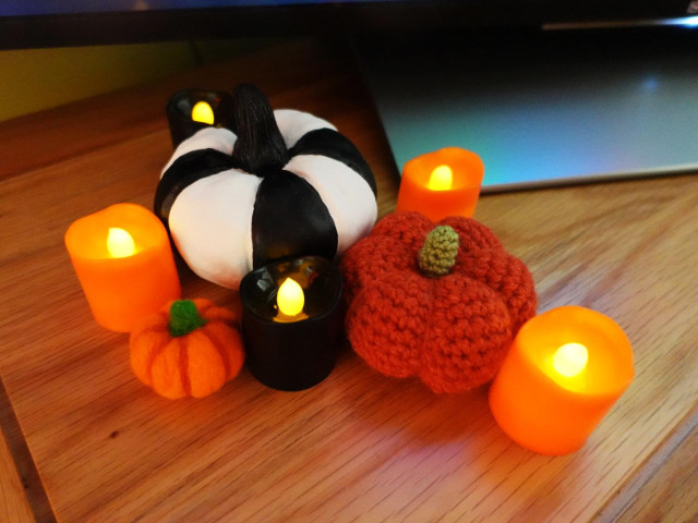 A group of three pumpkin ornaments, surrounded by small squat black and orange candles, sitting on a wooden surface. The pumpkins are all very different, the largest is made from air dry clay and painted in black and white stripes. The other two are both orange, one tiny needlefelted pumpkin and a crocheted one somewhere between the two sizes.