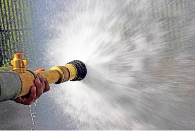 Photo: A hand in the foreground grasps a firehouse with a large yellow nozzle spraying a powerful jet of water throughout the rest of the frame.