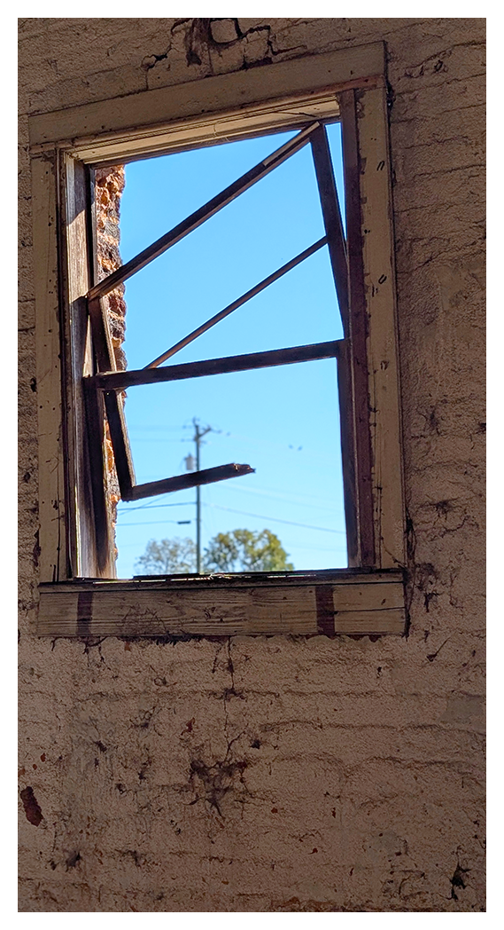 abandoned building interior. a damaged wood-framed window with no glass on a wall of old brick painted white at one time. power pole and lines and a couple trees under a clear blue sky are visible through the window. 