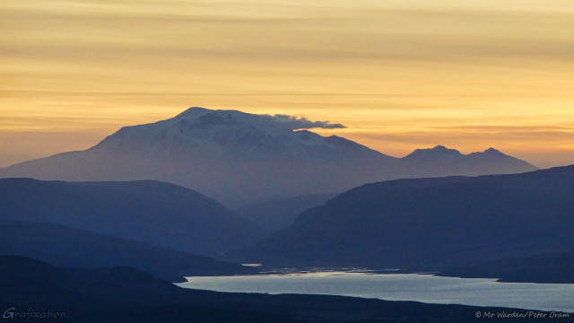 A photo of a mountain under a sky filled with streaked cloud. The sunset has tinted this in strong shades of ochre, orange and yellow. A beard of cloud is streaming from the top of the volcano, and close inspection reveals a snow cap and patches of ice down the flanks. The foreground is a large body of water, picking up a small reflection of the colours above, and the humped outlines of hills on either shore. These are in darkening shades of blue.