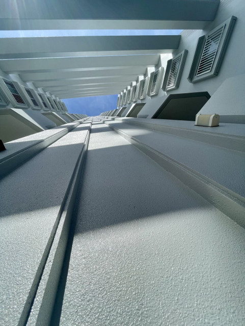 An upward view of a modern building's interior, showcasing white walls and slatted windows, under a bright blue sky. Sunlight creates a bright ambiance, emphasizing the architectural lines and design elements.