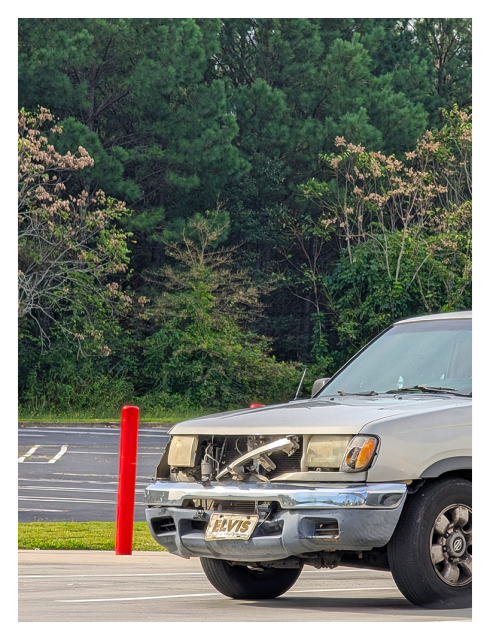 daytime. at the edge of the circle k parking lot. a small older pickup is backed into a parking space, the trucks front end has been banged up, but a bent-up ELVIS license plate is intact. a red parking bollard and strip of grass beside the truck. the background is an empty parking lot and woods bordering the other side.