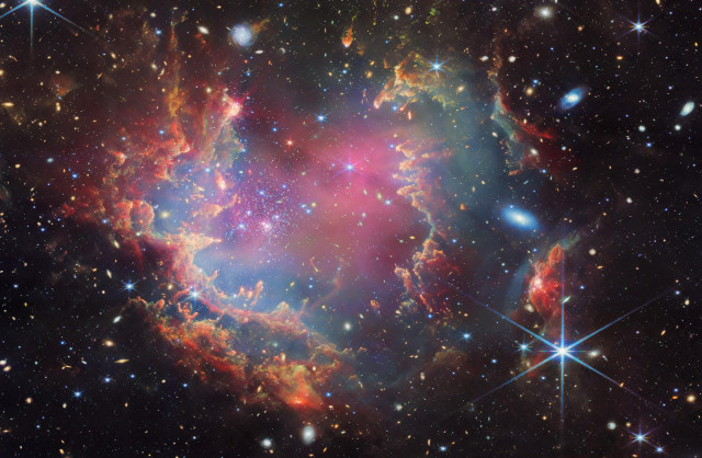 Near the outskirts of the Small Magellanic Cloud, a satellite galaxy roughly 200 000 light-years from Earth, lies the young star cluster NGC 602, which is featured in this new image from the NASA/ESA/CSA James Webb Space Telescope. This image includes data from Webb’s NIRCam (Near-InfraRed Camera) and MIRI (Mid-InfraRed Instrument).

The local environment of this cluster is a close analogue of what existed in the early Universe, with very low abundances of elements heavier than hydrogen and helium. The existence of dark clouds of dense dust and the fact that the cluster is rich in ionised gas also suggest the presence of ongoing star formation processes. This cluster provides a valuable opportunity to examine star formation scenarios under dramatically different conditions from those in the solar neighbourhood.