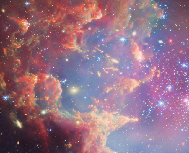 Near the outskirts of the Small Magellanic Cloud, a satellite galaxy roughly 200 000 light-years from Earth, lies the young star cluster NGC 602, which is featured in this new image from the NASA/ESA/CSA James Webb Space Telescope. This image includes data from Webb’s NIRCam (Near-InfraRed Camera) and MIRI (Mid-InfraRed Instrument).

The local environment of this cluster is a close analogue of what existed in the early Universe, with very low abundances of elements heavier than hydrogen and helium. The existence of dark clouds of dense dust and the fact that the cluster is rich in ionised gas also suggest the presence of ongoing star formation processes. This cluster provides a valuable opportunity to examine star formation scenarios under dramatically different conditions from those in the solar neighbourhood.