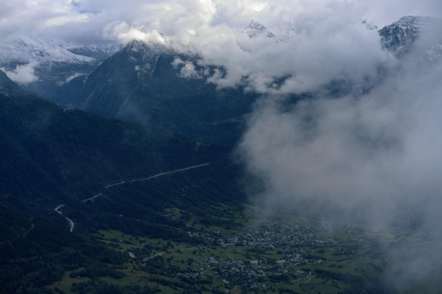 An aerial view of a small Alpine village nestled in a dramatic mountain valley. Snow-capped peaks pierce through clouds at the top of the frame, while a winding road cuts through the forested mountainside. The village sits in a patch of green meadows at the valley floor, partially obscured by rolling clouds. The scene captures the impressive scale of the Alps, with the tiny settlement dwarfed by the surrounding mountains.