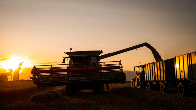 Ein Mähdrescher schüttet seine Ernte in einen daneben stehenden Anhänger, der von einem Traktor gezogen wird.
Links ist ein Kraftwerk zu sehen, über dem die Sonne untergeht. Die ganze Szene wird dadurch in goldenes Licht getaucht. 