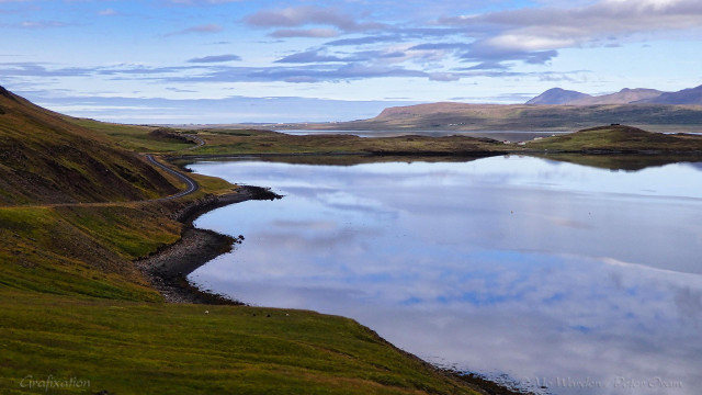 A photo of a landscape containing a large body of water. The shores are green with vegetation and, on the nearest bank on the left of the shot, sheep can be seen lying in the grasses. A road winds away from left centre and towards the horizon. A promontory juts into the water and a few cottages are nestled upon it. The sky is summer cyan except for a few thin clouds. The water surface is reflecting this, apart from a small area where a breeze has ruffled the surface. Nearby, dots of birds can just be seen, and the float of a crab pot.