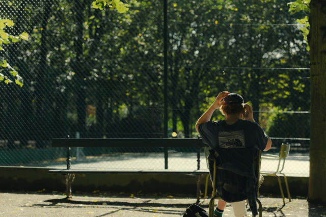 A person wearing a black North Face t-shirt and baseball cap sits alone on a green chair, photographed from behind. They're adjusting their cap while seated in what appears to be a park or outdoor recreational area with a chain-link fence and trees in the background. The scene is bathed in warm afternoon sunlight, casting long shadows on the ground.