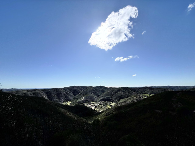 A panoramic view of rolling green hills under a clear blue sky with a single fluffy cloud. Sunlight highlights the contours of the landscape, creating a serene and picturesque scene.