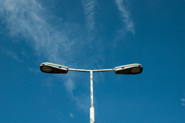 Photo of the top of a tatty white double-headed lamppost against a bright blue sky with streaks of cloud, possibly from dissolving contrails.