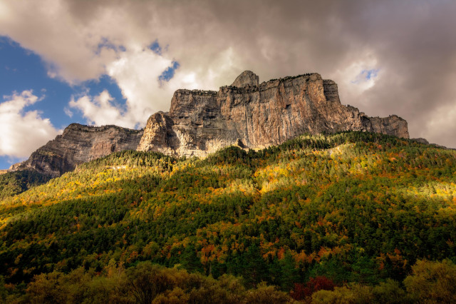 Fotografía donde se puede en la mitad inferior de la misma se puede ver una gran falda de montaña llena de árboles con colores de otoño y en la superior una formación montañosa con nubes por encima.