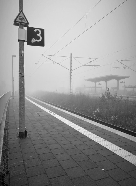 Motiv:
Ein menschenleerer Bahnsteig in dichtem Nebel. Die Nebelstimmung lässt den Hintergrund verschwinden und verstärkt das Gefühl von Einsamkeit und Isolation. Die Gleise, das Wartehäuschen und die Oberleitungen laufen in der Ferne in den Nebel hinein, was der Szene eine fast endlose Tiefe verleiht.

Bildformat:
Hochformat.

Genre:
Urbaner Minimalismus und Melancholie. Das Bild transportiert eine stille und einsame Atmosphäre, wie sie oft in frühen Morgenstunden zu spüren ist.

Farben:
Schwarz-Weiß-Bild, das die neblige Szenerie verstärkt und der Aufnahme eine zeitlose Stimmung verleiht.

Lichtsituation:
Weiches, diffuses Licht, das vom Nebel gestreut wird, ohne klare Schatten oder harte Kontraste. Die Beleuchtung unterstützt die Ruhe und Stille des Moments und verstärkt die Melancholie der Szene.