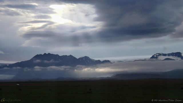 A photo of mountains, with the top of a wide glacier visible between them. The foreground is a dark surface, and a car can be seen travelling into the frame from the left. The sky is overcast and some mist has gathered over the lower slopes, hiding large parts of the glacier tongues. Just left of top centre, a window has opened in the cloud and sunbeams are streaming through to illuminate the fog below.