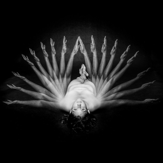 A naked woman is lying on a dark background with her face towards the camera. Her arms start outstretched on her side in the first exposure and gradually shift up along the nine exposures of the shot. Black and white. 