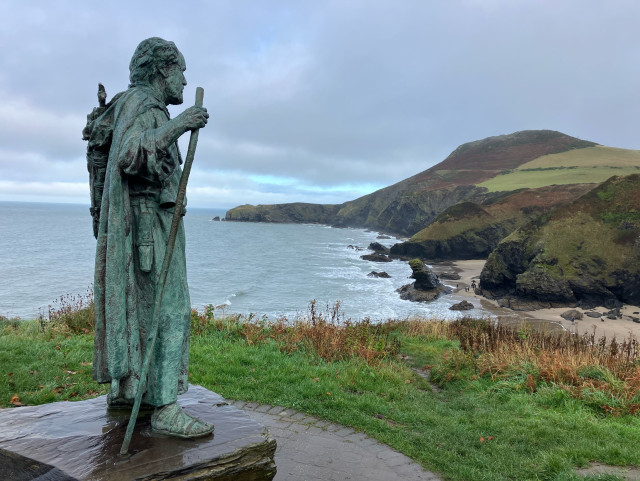 A photo of the bronze statue of Saint Carannog standing, with his staff, on cliffs overlooking Llangrannog beach. The Lochtyn peninsula ahead along the coast - a high bracken covered hill that slopes down to a thin, finger-like promontory. Rugged cliffs and sandy coves in the mid distance. The sky is thick with grey cloud but there is a hint of blue over the peninsula.