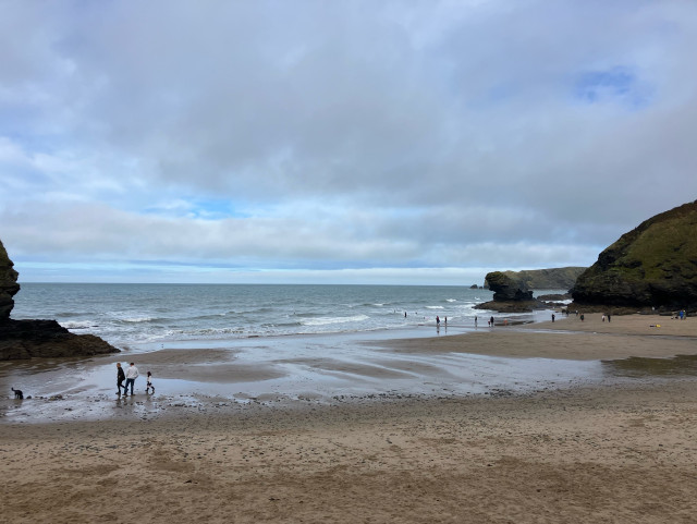 Photo of a sandy beach at low ish tide. A few tiny figures of people and dogs on the sand and surfers in the waves. A rock stack to the right - Carreg Bica, said to be the tooth of a giant. The sky is cloudy with patches of blue opening.