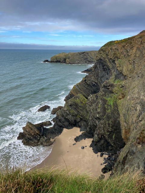 A photo of the peninsula from further along the coast path, a sandy beach below dark rocky cliffs. 
There is a patch of turquoise blue sky that appears sandwiched between grey cloud. The sea is a grey blue with white waves breaking against the base of the cliffs and coves.