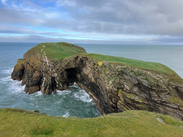 Photo of a small rocky island at the tip of the peninsula. Can’t quite get it all in frame, at least not without getting uncomfortably close to the cliff edge. The top of the island is turfed with short grass. The sheer cliffs show an interesting geology of folds. White streaks through the dark grey, towards the tip on the left, I’m guessing are intrusions of a different rock. A dark sea cave in the centre. The sky is cloudy with blue patches.