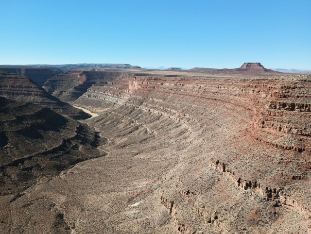 Color landscape photo of a deep canyon with a muddy looking river at the bottom. There are many layers of rock forming step like terraces as the canyon cliff descends to the river. The sky is blue. A small flat top mesa is seen on the right side of the frame a short distance from the canyon rim.