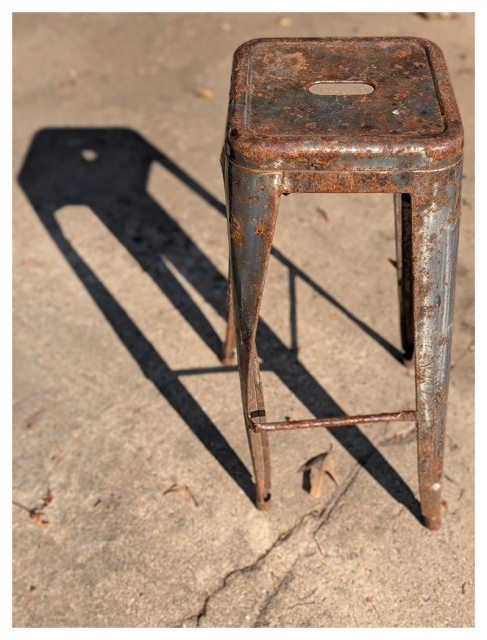 afternoon sunlight. a vintage metal stool with vent hole on the seat stands on cracked concrete. it casts a shadow to the upper left, onto concrete scatter with fallen leaves and other detritus. a sundial.