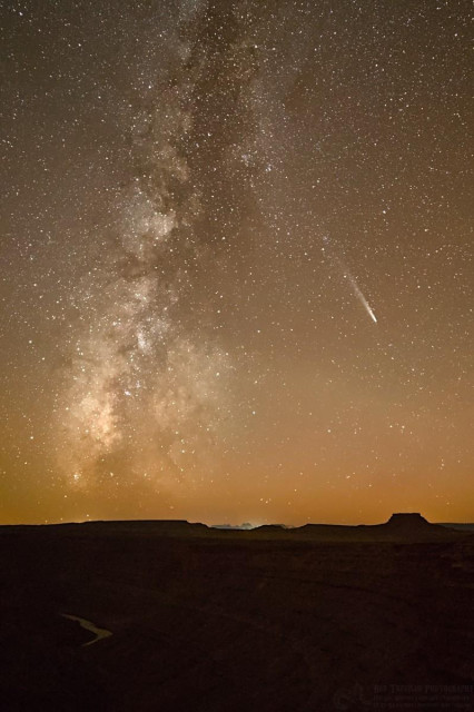 A color portrait photo showing the Milky Way galaxy which appears as a fuzzy white streak from the lower left to upper right of center. It kind of looks like a caterpillar with a dark segmented body and bright white legs. Mid-frame and to the right of the Milky Way is a comet, a small bright blob with a long fuzzy tail. The comet is fairly small. At the base of the photo and to the left, is a ribbon looking river. The horizon features squared off mesas.  