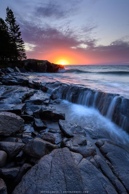 A photo of the sun rising over Lake Superior. In the background is the horizon and a fur tree on the left. In the foreground is a triangle shaped rock. There's a one foot ledge and the waves of Lake Superior are spilling over it. The overall color is blue/purple with orange and yellow in the sun and across the wet rocks.