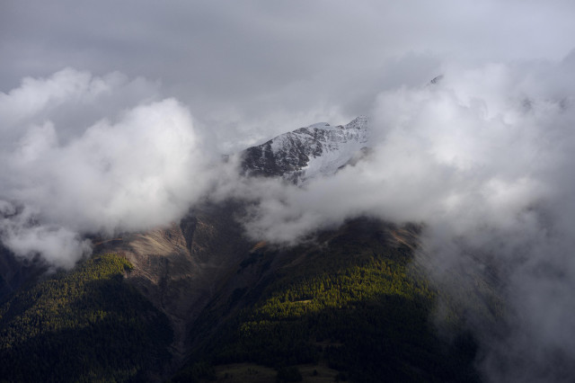 A mountainous landscape where dramatic white clouds partially shroud a snow-capped peak. The mountain slopes transition from snow at the summit through rocky terrain to forested areas with evergreen trees at lower elevations. The scene has a moody atmosphere with the interplay of clouds, snow, and dark mountain features.