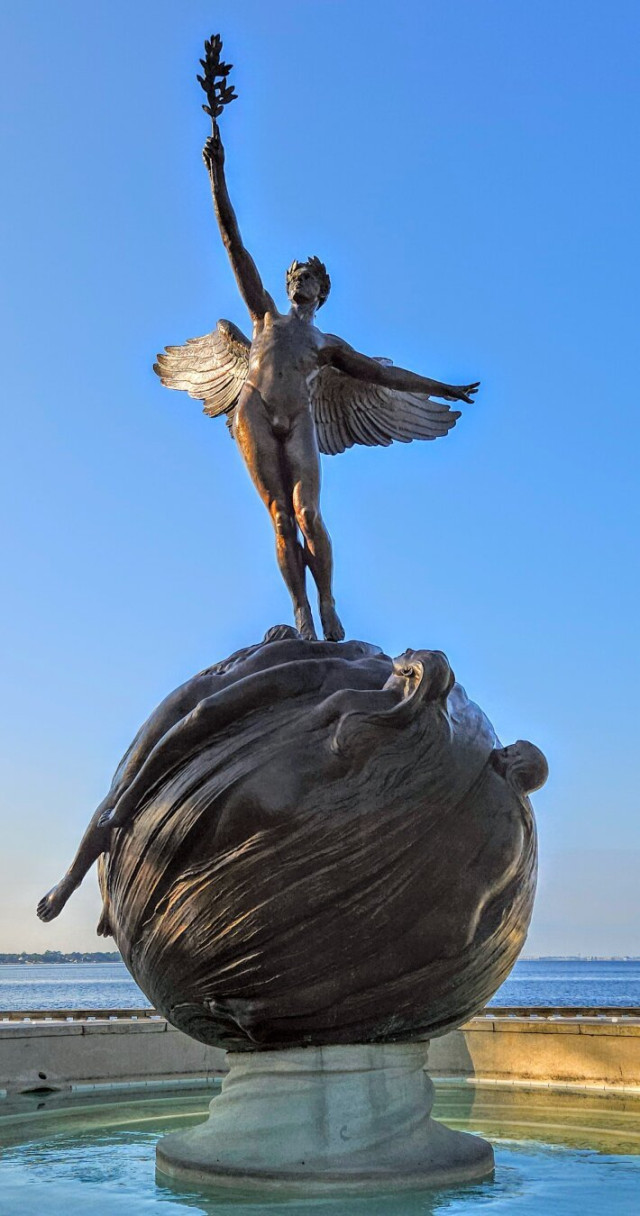 Close up view of the sculpture of an angelic figure holding an olive branch, as if an offering, high into the sky above. He is standing on a globe within a city park fountain. With a vast blue river beyond, all below a big, clear blue Florida sky.