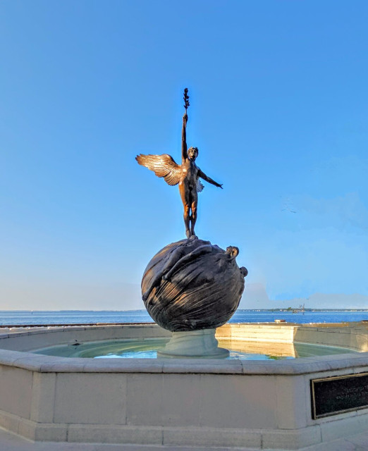 Distant view of the sculpture of an angelic figure holding an olive branch, as if an offering, high into the sky above. He is standing on a globe within a city park fountain. With a vast blue river beyond, all below a big, clear blue Florida sky.