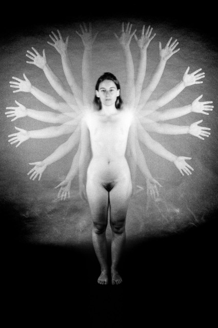 A naked woman is standing in front of the camera, with 9 pairs of arms outstretched around her. Black and white. 